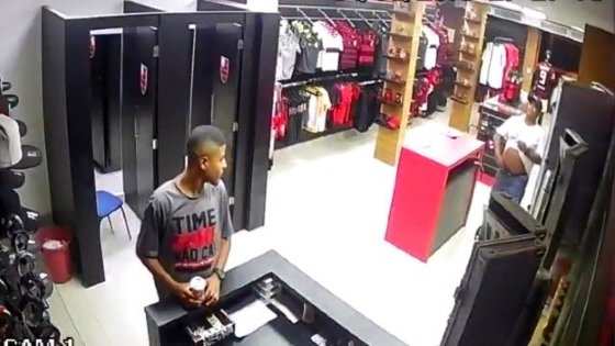 Um dos assaltantes, de camisa branca, mostra a arma, anuncia o roubo e rende funcionários da loja